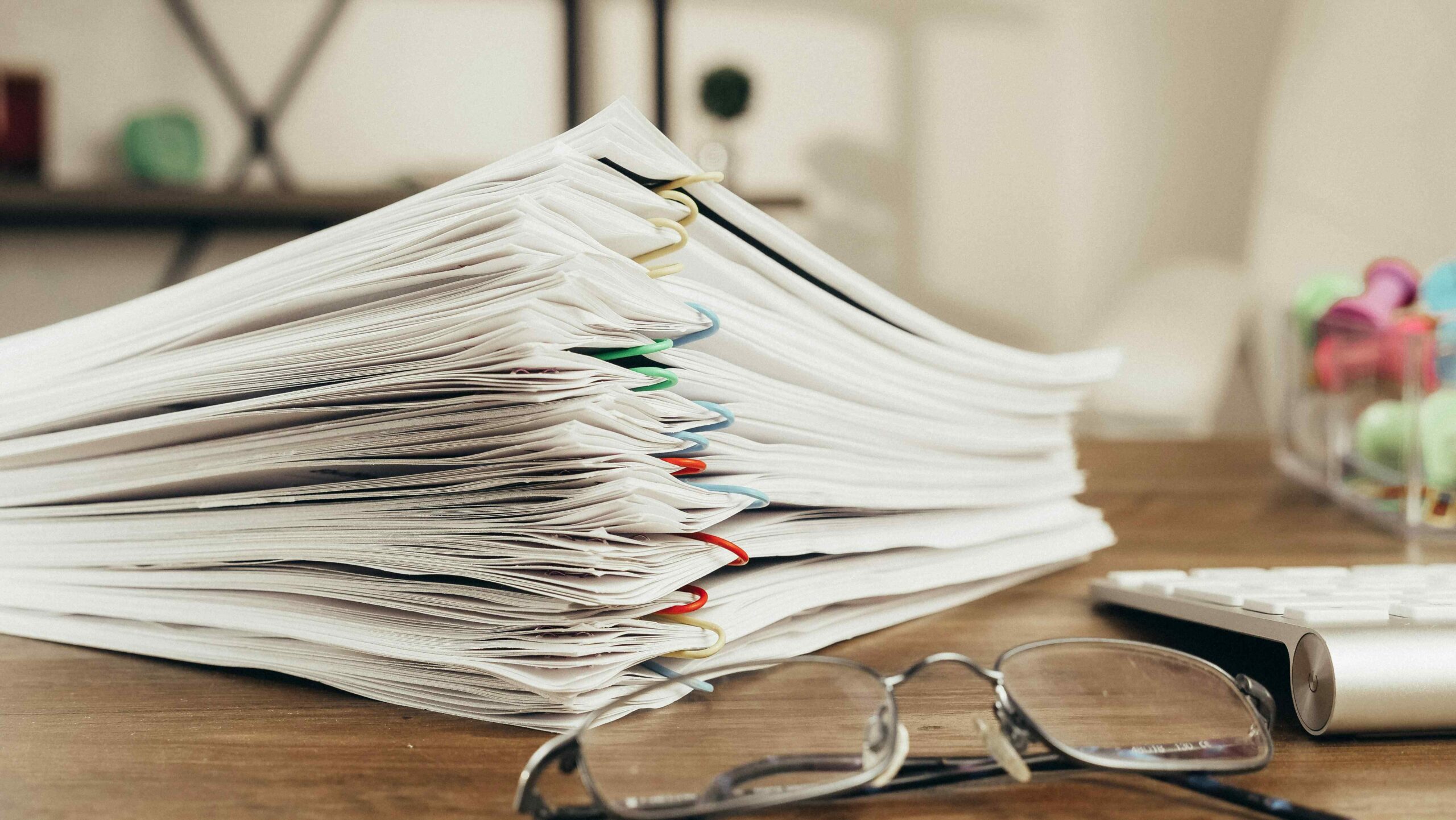 Pliki papieru leżące na stole obok okularów i innych artykułów biurowych.